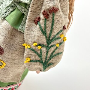 Puede incluir: Una mu&ntilde;eca hecha a mano con un corpi&ntilde;o verde y mangas de tela de espiga beige. Las mangas est&aacute;n bordadas con tallos verdes y flores marrones y amarillas. La mu&ntilde;eca tiene pelo largo casta&ntilde;o y est&aacute; adornada con una corona floral.