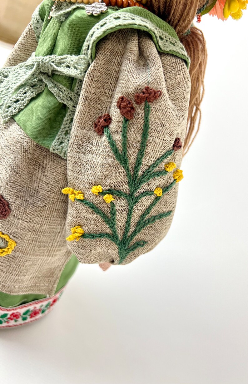 Puede incluir: Una mu&ntilde;eca hecha a mano con un corpi&ntilde;o verde y mangas de tela de espiga beige. Las mangas est&aacute;n bordadas con tallos verdes y flores marrones y amarillas. La mu&ntilde;eca tiene pelo largo casta&ntilde;o y est&aacute; adornada con una corona floral.