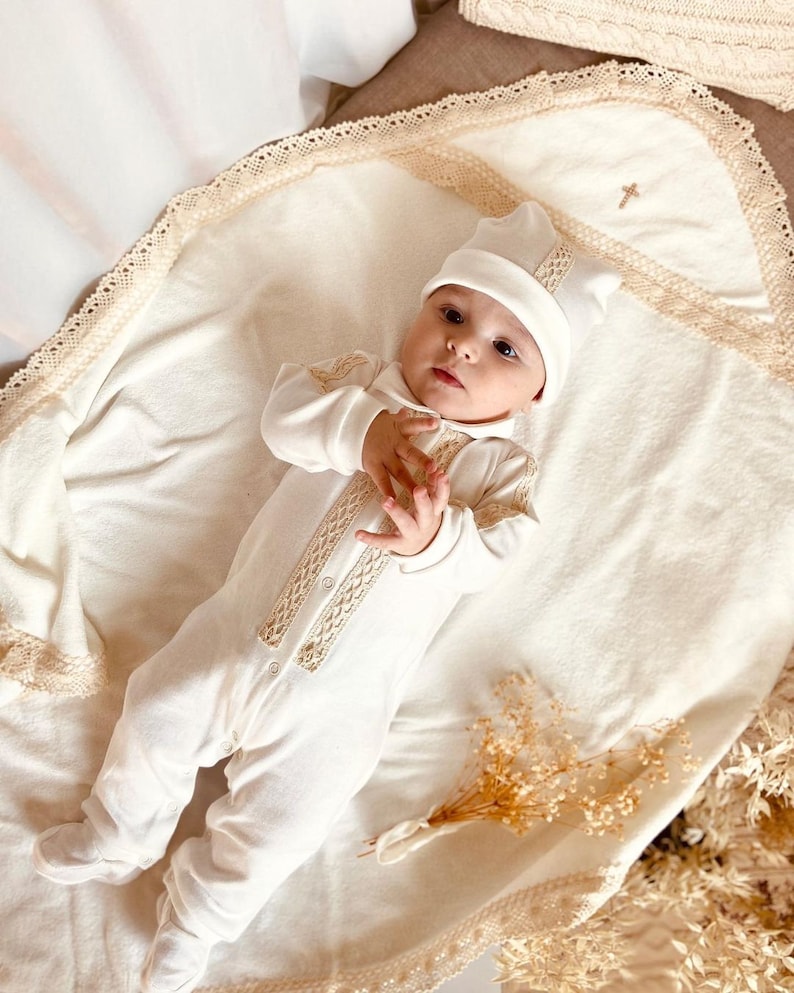 Op de afbeelding: Een witte baby-outfit met een kantversiering en een bijpassende muts. De outfit ligt op een witte deken met een kantversiering. De baby draagt een witte muts met een kantversiering.