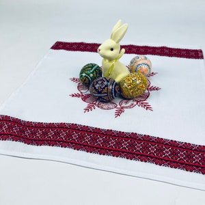 May include: White embroidered linen table runner with a red border. A white ceramic bunny sits on the runner surrounded by five colorful Easter eggs.