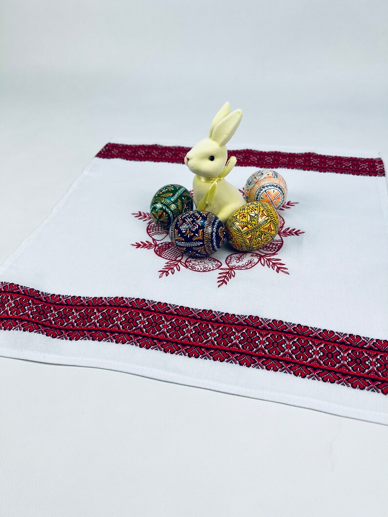 May include: White embroidered linen table runner with a red border. A white ceramic bunny sits on the runner surrounded by five colorful Easter eggs.
