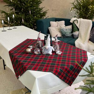 May include: A festive Christmas table setting with a red and green plaid table runner. The table is decorated with gnome figurines, ornaments, and a clear glass globe. Christmas trees and gifts are in the background.