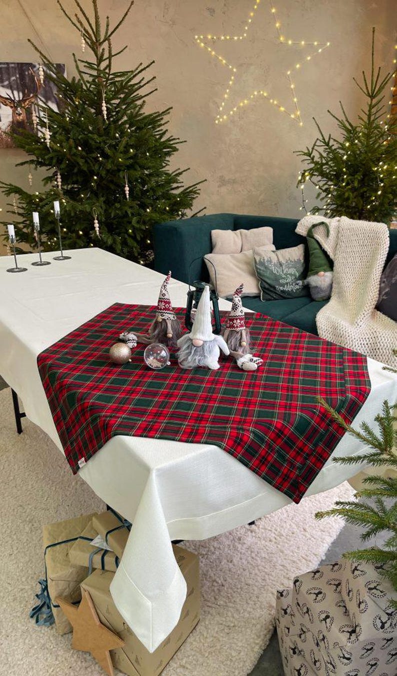 May include: A festive Christmas table setting with a red and green plaid table runner. The table is decorated with gnome figurines, ornaments, and a clear glass globe. Christmas trees and gifts are in the background.