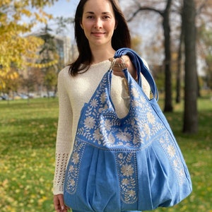 Peut inclure: Un grand sac cabas bleu avec des motifs floraux brod&eacute;s blancs. Le sac a un fond pliss&eacute; et une seule bandouli&egrave;re.