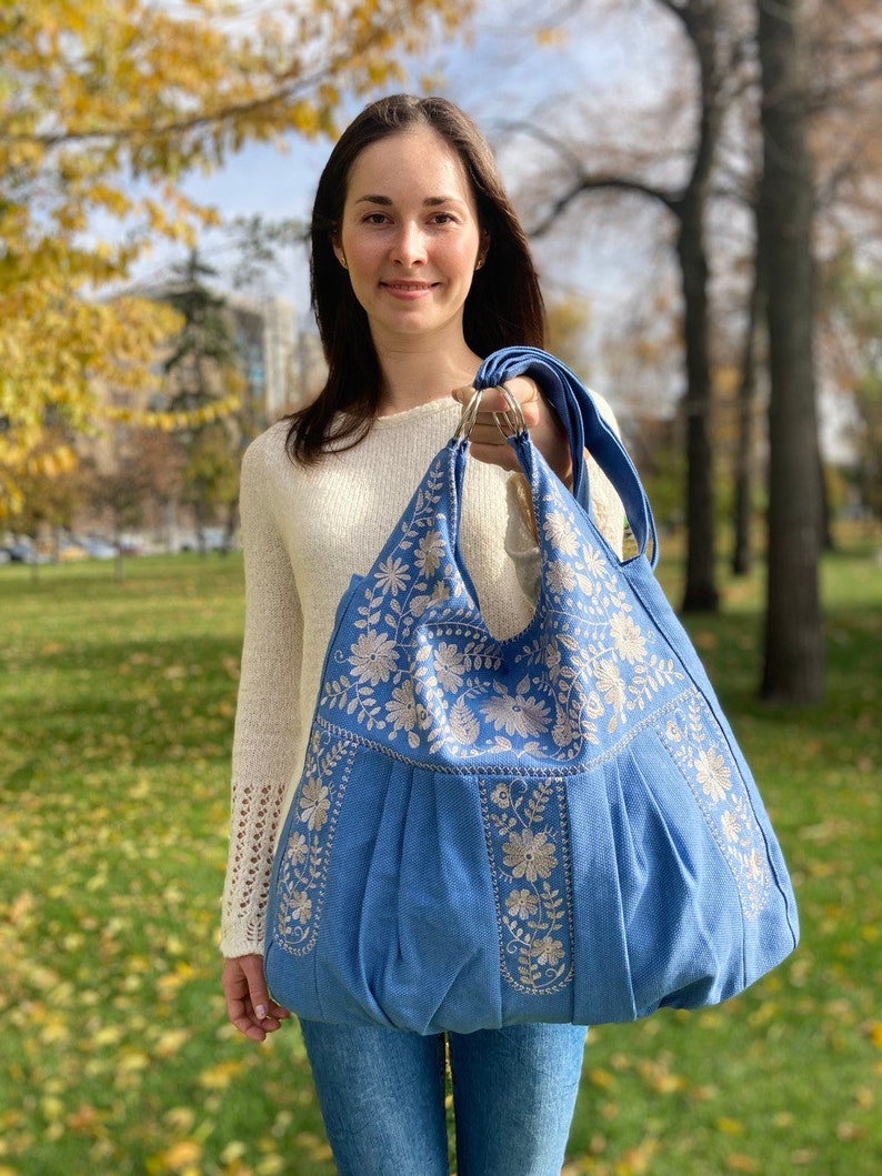 Peut inclure: Un grand sac cabas bleu avec des motifs floraux brod&eacute;s blancs. Le sac a un fond pliss&eacute; et une seule bandouli&egrave;re.