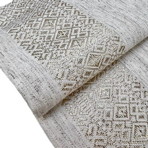 Puede incluir: Textil tejido blanco roto doblado con un patr&oacute;n geom&eacute;trico de diamantes en un tono m&aacute;s claro. La tela tiene una apariencia texturizada, con la secci&oacute;n estampada a lo largo del borde.