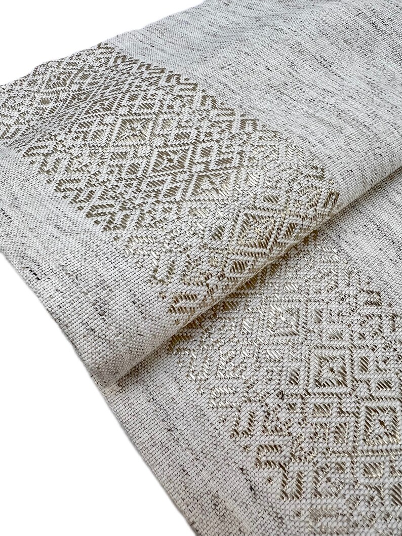 Puede incluir: Textil tejido blanco roto doblado con un patr&oacute;n geom&eacute;trico de diamantes en un tono m&aacute;s claro. La tela tiene una apariencia texturizada, con la secci&oacute;n estampada a lo largo del borde.