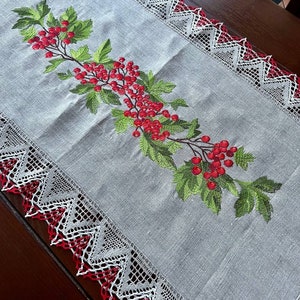 May include: A white linen table runner with a red and white lace border. The runner features an embroidered design of a green branch with red berries.
