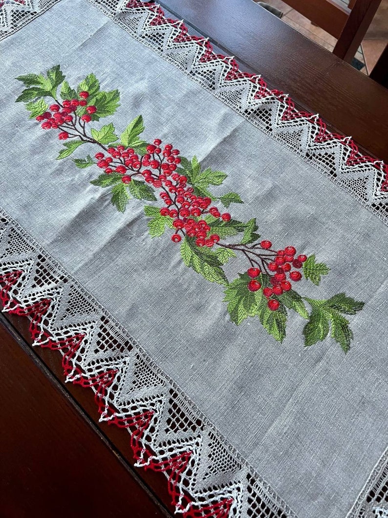 May include: A white linen table runner with a red and white lace border. The runner features an embroidered design of a green branch with red berries.