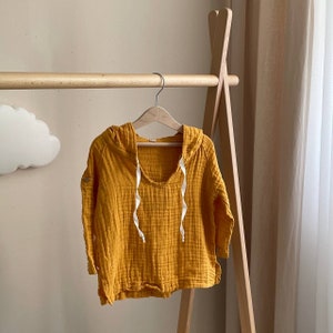 May include: A mustard yellow hooded sweatshirt with a white drawstring. The sweatshirt is made of a soft, textured fabric and has a relaxed fit.