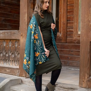 May include: A woman wearing a dark green dress and a teal shawl with orange floral patterns. She is wearing black tights and olive green heeled boots. The background is a wooden building with a door and window.