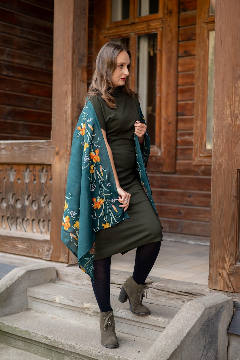 May include: A woman wearing a dark green dress and a teal shawl with orange floral patterns. She is wearing black tights and olive green heeled boots. The background is a wooden building with a door and window.