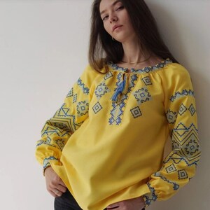 May include: A yellow embroidered blouse with blue geometric patterns. The blouse has a round neckline and long sleeves.
