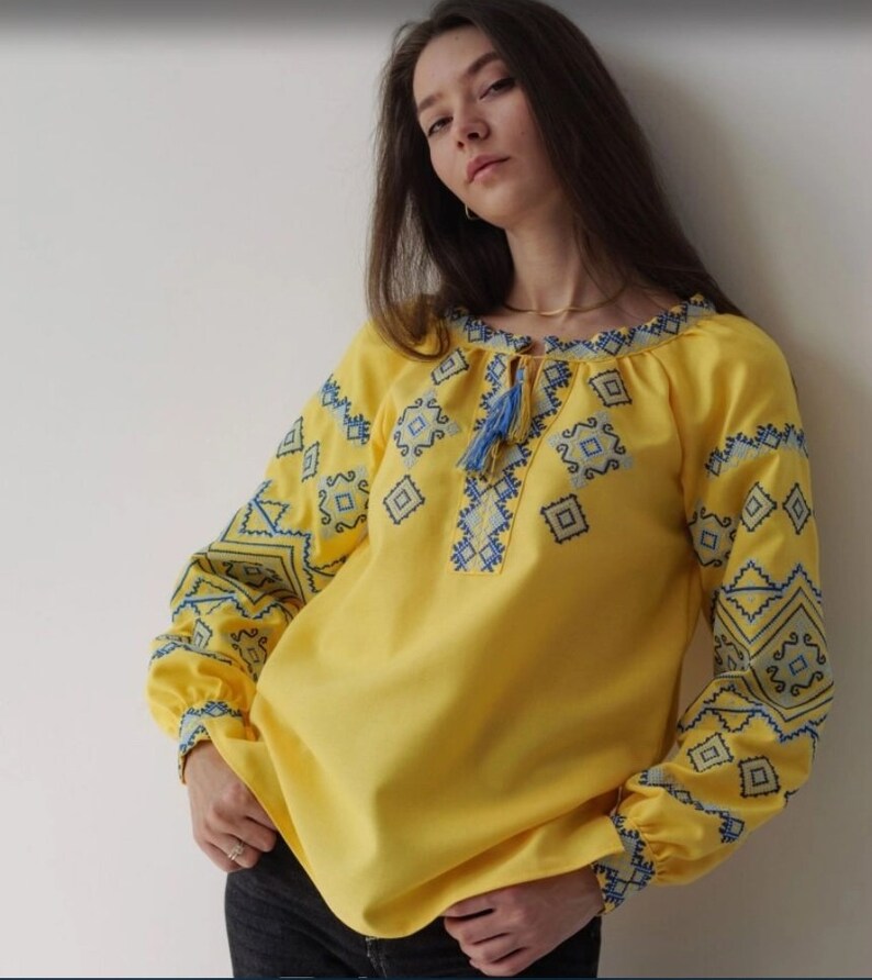 May include: A yellow embroidered blouse with blue geometric patterns. The blouse has a round neckline and long sleeves.