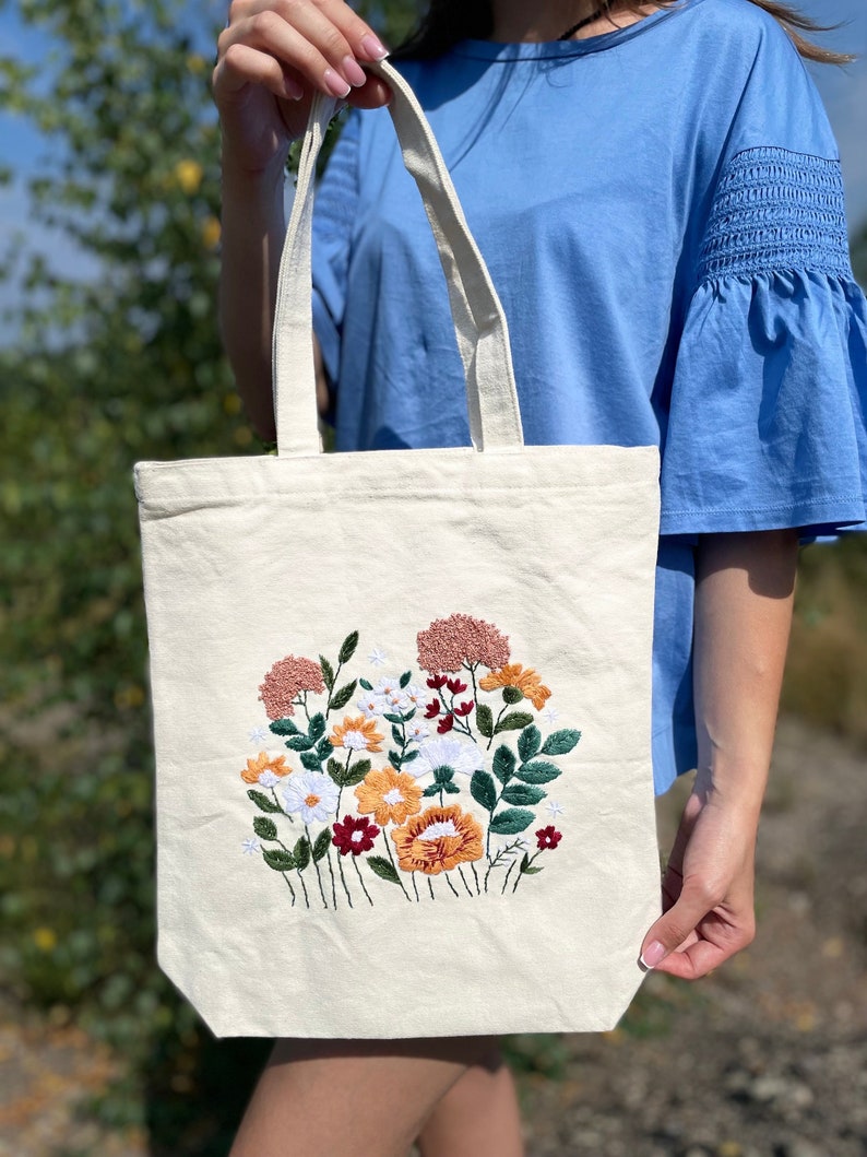 May include: A white canvas tote bag with a floral embroidery design. The embroidery features a variety of flowers in shades of pink, red, orange, yellow, and green.