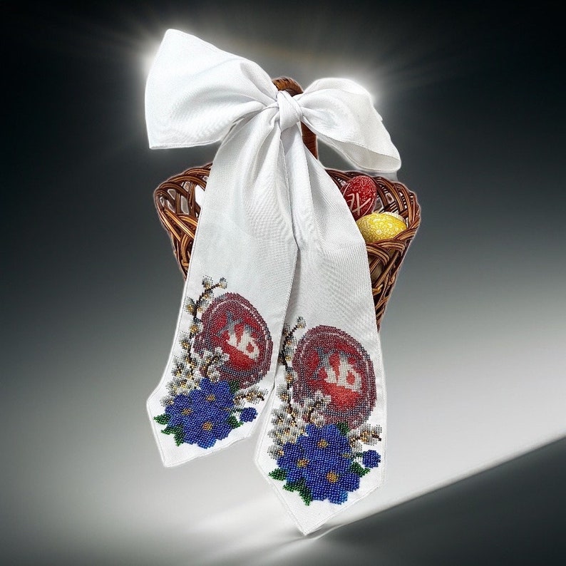 May include: A woven basket filled with decorated Easter eggs, adorned with a large white bow. Two white fabric panels hang from the basket, each featuring a red and white embroidered design with blue floral accents.