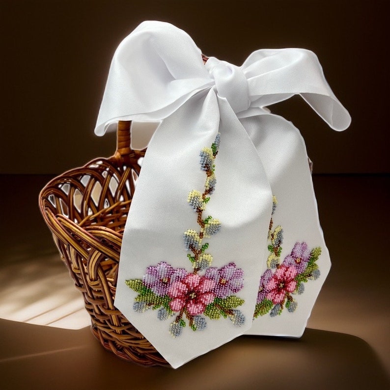 K&ouml;nnte beinhalten: Ein wei&szlig;es Satinband mit Schleife, bestickt mit zarten Blumendesigns in Rosa-, Lila- und Gr&uuml;nt&ouml;nen. Das Band ist um einen Weidenkorb gebunden.