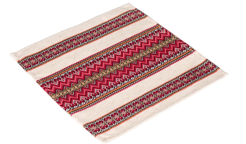 Op de afbeelding: Cr&egrave;mekleurig vierkant textiel met een gestreept ontwerp. De strepen hebben ingewikkelde rode en gouden geometrische patronen, waardoor een levendig contrast ontstaat met de neutrale achtergrond. Het textiel lijkt van geweven stof te zijn.