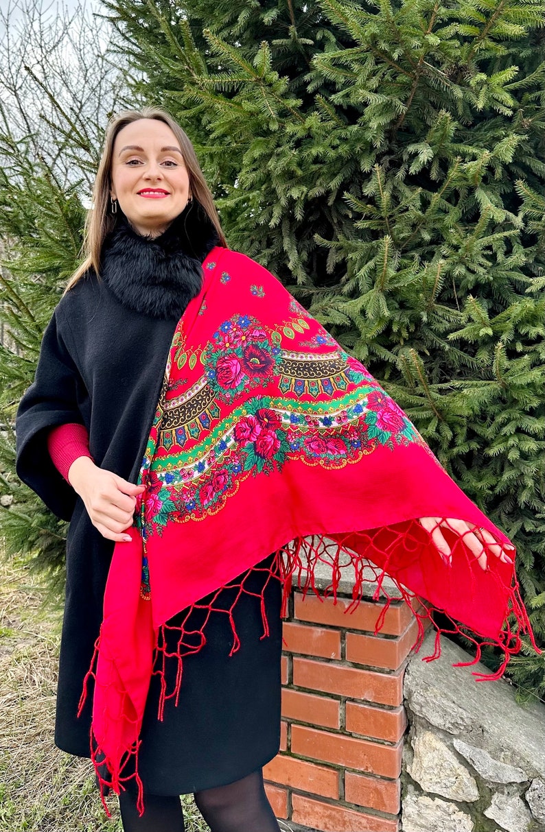 May include: A woman wearing a black coat with a fur collar and a red shawl with floral embroidery and fringe. The shawl is draped over her shoulders and has a traditional design.