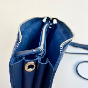 May include: A navy blue leather crossbody bag with two zippered compartments and a silver metal grommet. The bag is open and the lining is visible.