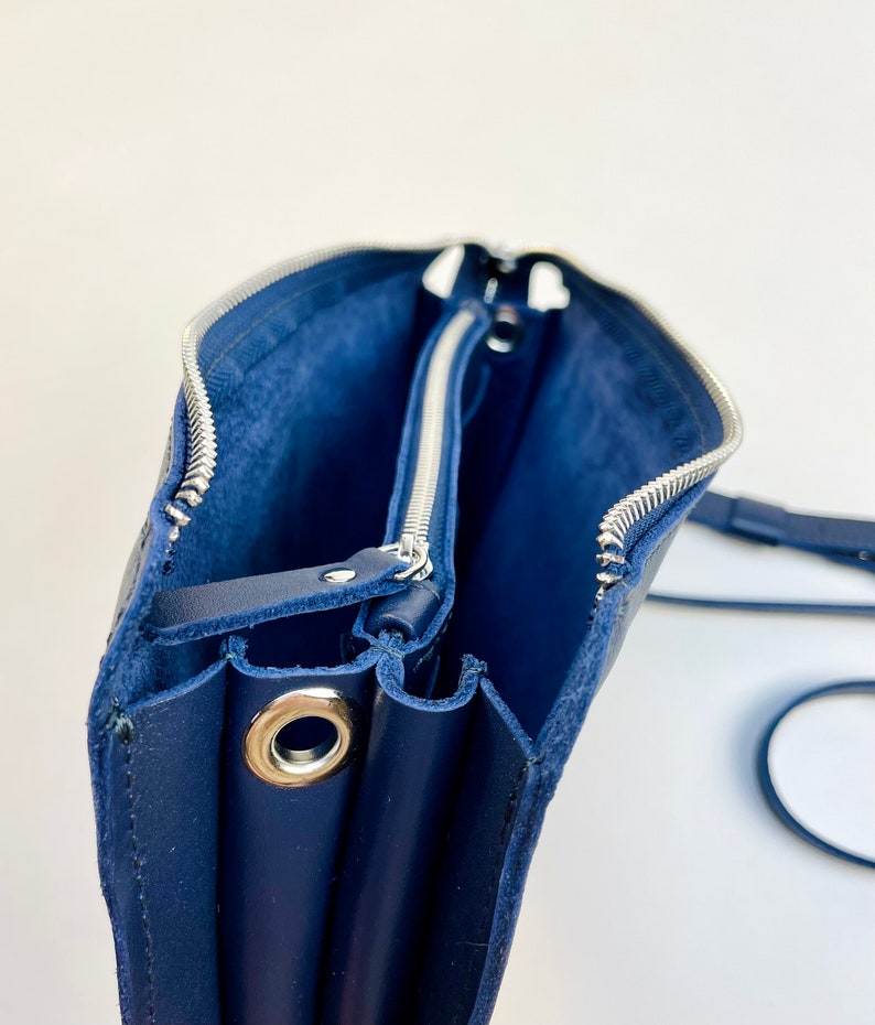 May include: A navy blue leather crossbody bag with two zippered compartments and a silver metal grommet. The bag is open and the lining is visible.
