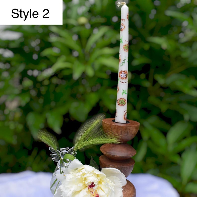 May include: A white candle with a decorative pattern of circular designs and green leaves, set in a wooden candle holder. The candle holder is made of stacked, rounded wooden pieces. The text "Style 2" is in the upper left corner.
