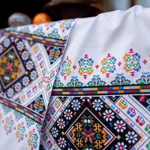 May include: White fabric with colorful embroidered floral patterns. The embroidery is in a geometric design with black, red, blue, green, yellow, and orange thread.