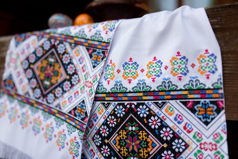 May include: White fabric with colorful embroidered floral patterns. The embroidery is in a geometric design with black, red, blue, green, yellow, and orange thread.
