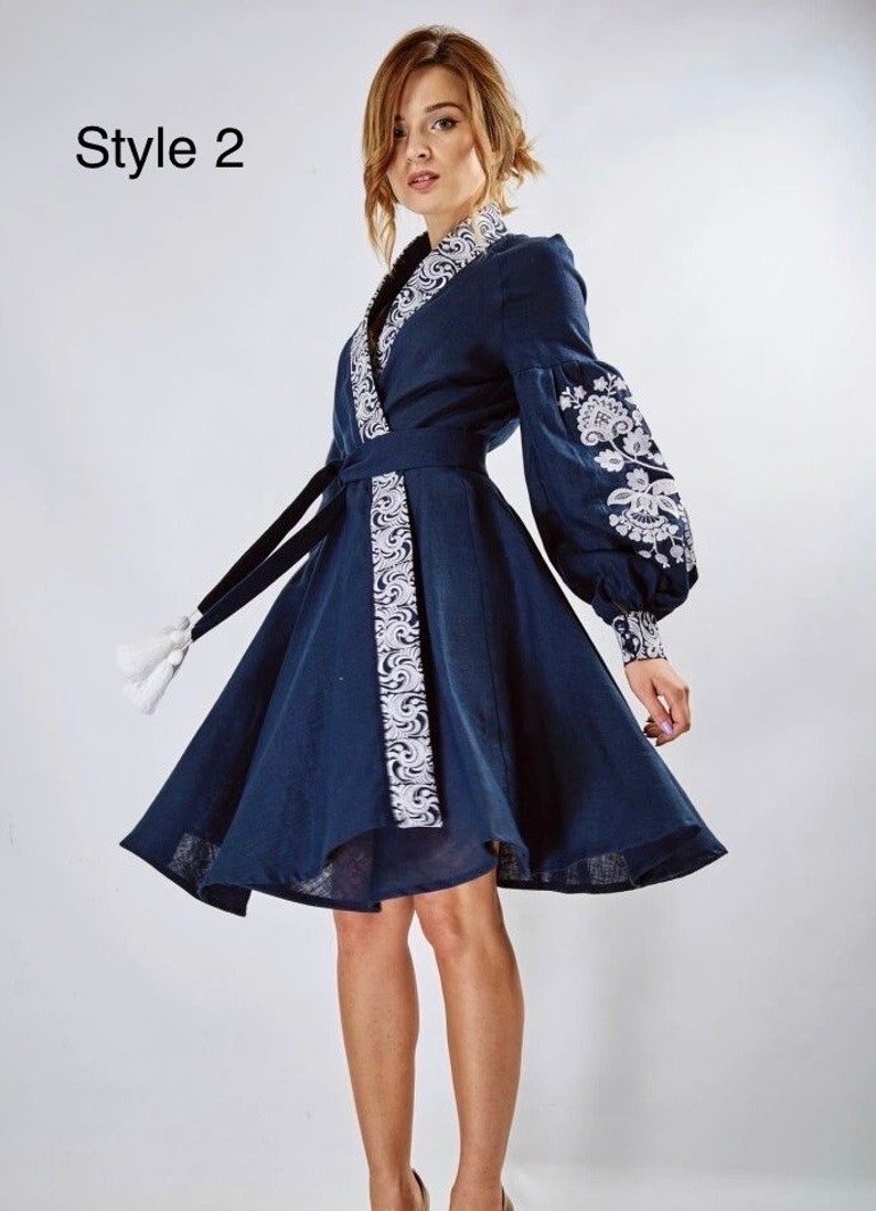 May include: A navy blue linen dress with white embroidery. The dress has a wrap style and a belt with white embroidery. The dress has long sleeves with white embroidery on the cuffs. The dress has a flared skirt. The dress is labelled "Style 2".