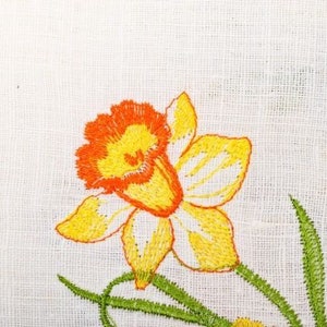 Puede incluir: Flor bordada de color amarillo y naranja con tallos verdes sobre una tela de lino blanca.