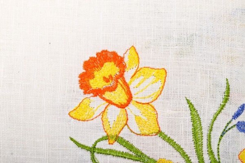 Puede incluir: Flor bordada de color amarillo y naranja con tallos verdes sobre una tela de lino blanca.