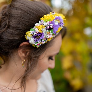 May include: A woman wearing a white embroidered blouse with a red accent and a floral headband with yellow, purple, and white flowers.