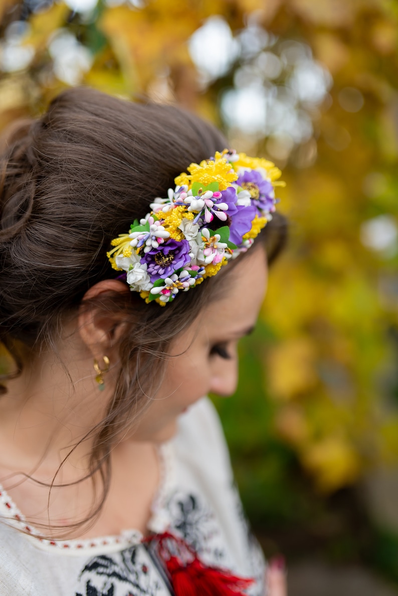 May include: A woman wearing a white embroidered blouse with a red accent and a floral headband with yellow, purple, and white flowers.