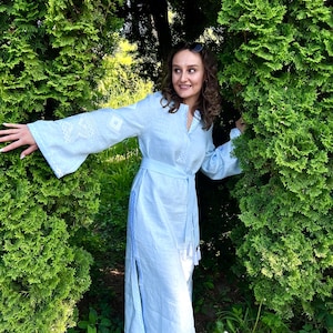 Pode incluir: Um vestido de linho azul claro com detalhes bordados brancos nas mangas e no corpete. O vestido apresenta decote em V, cinto a condizer e fenda lateral. A mulher usa sand&aacute;lias pretas e &oacute;culos de sol.