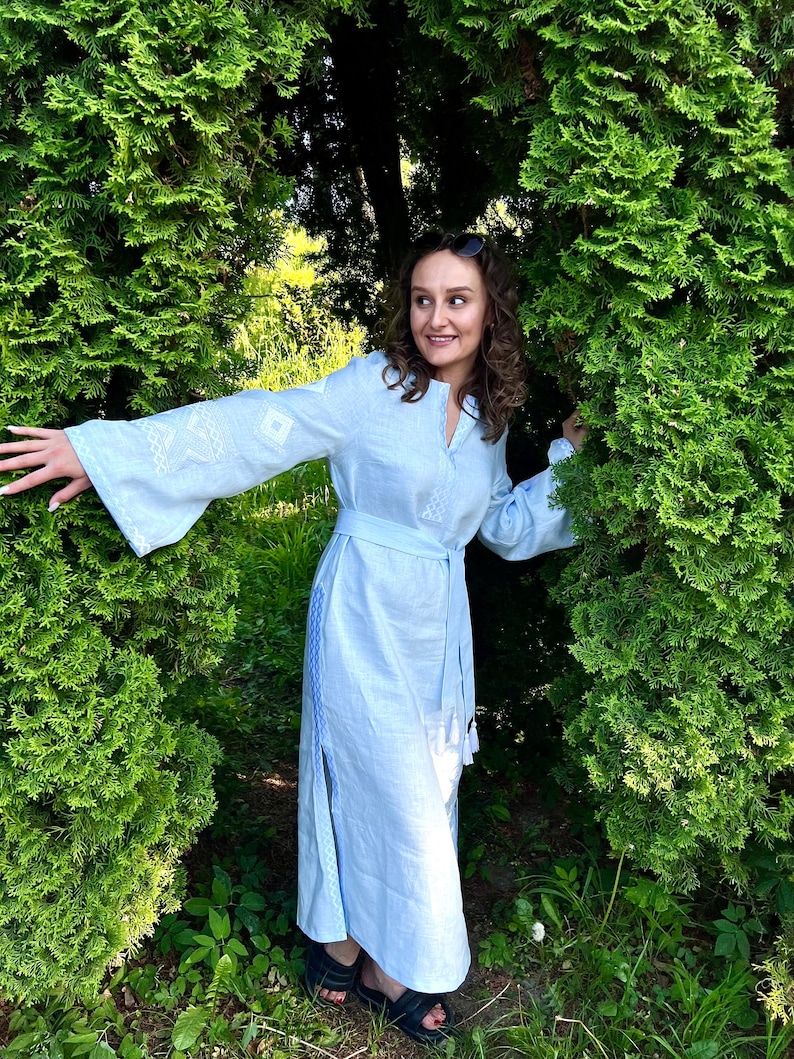 Pode incluir: Um vestido de linho azul claro com detalhes bordados brancos nas mangas e no corpete. O vestido apresenta decote em V, cinto a condizer e fenda lateral. A mulher usa sand&aacute;lias pretas e &oacute;culos de sol.