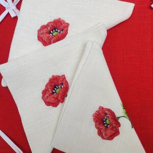 May include: Three white linen napkins with red embroidered poppy flowers. The napkins are folded in a triangular shape and are arranged on a red background.