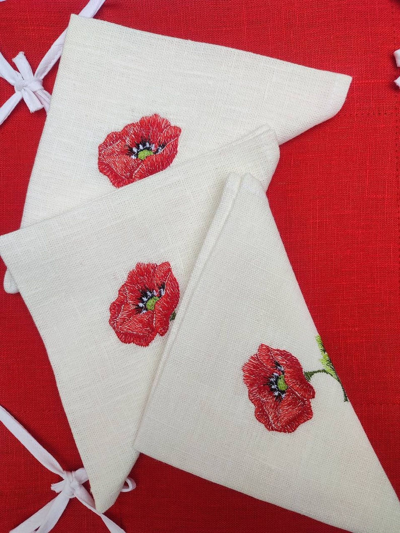 May include: Three white linen napkins with red embroidered poppy flowers. The napkins are folded in a triangular shape and are arranged on a red background.
