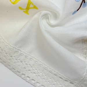 May include: White linen fabric with embroidered floral and letter details. The embroidery features pink, blue, and green flowers, and yellow letters. A delicate white lace trim borders the edge of the fabric.