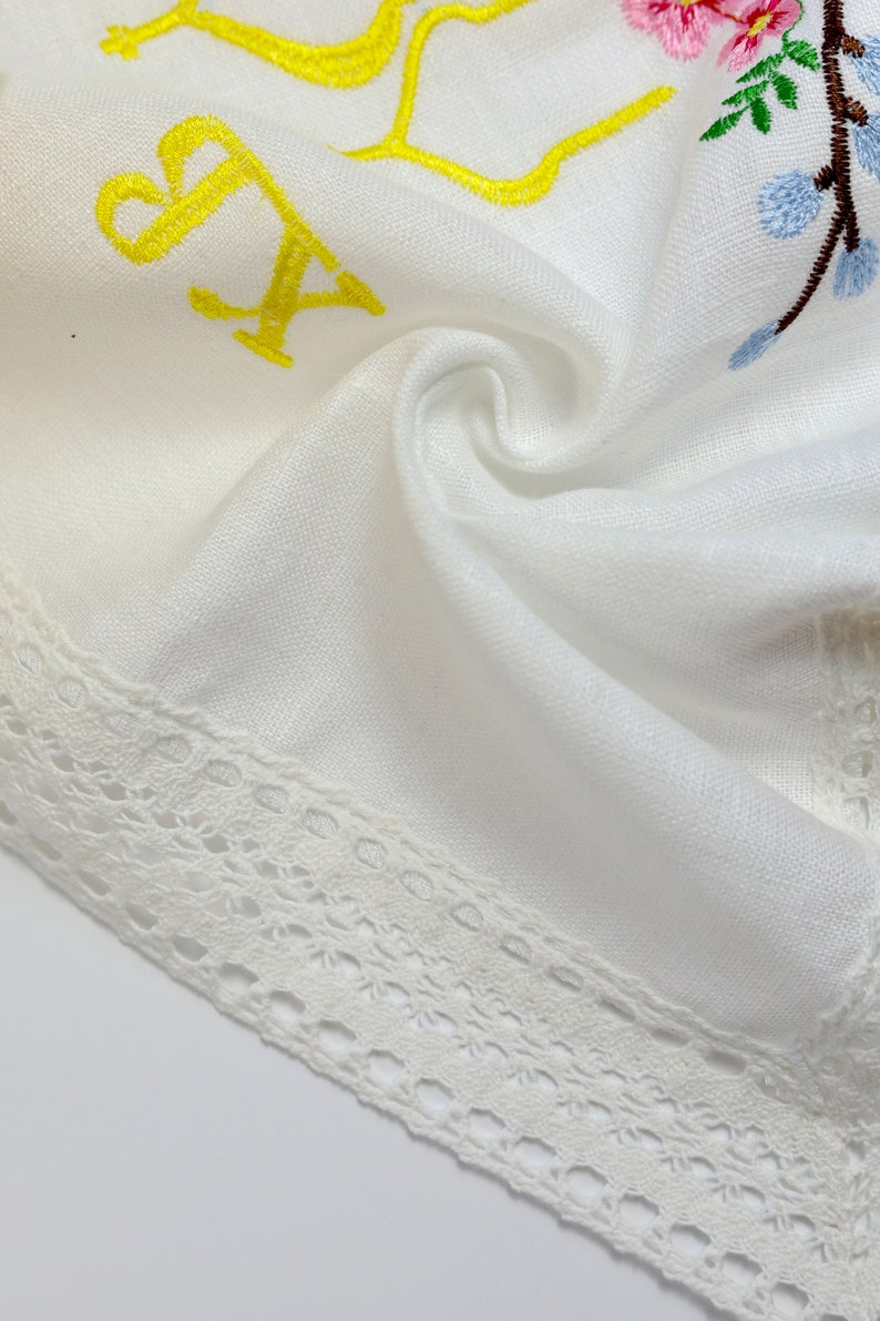 May include: White linen fabric with embroidered floral and letter details. The embroidery features pink, blue, and green flowers, and yellow letters. A delicate white lace trim borders the edge of the fabric.