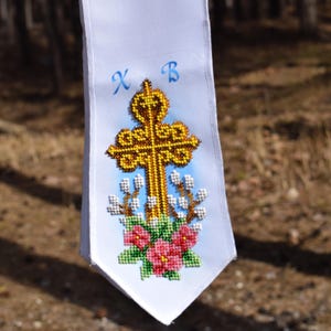 May include: White fabric with a cross-stitch design featuring a gold cross, pink flowers, and blue lettering. The cross is adorned with intricate beadwork, and the flowers are in full bloom. The background has a light blue gradient.