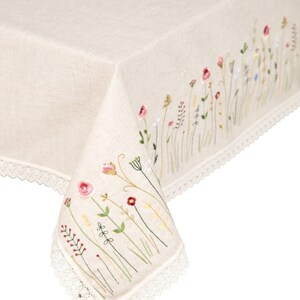 May include: A cream-colored linen tablecloth with embroidered wildflowers in pink, red, yellow, and green. The edge is finished with a delicate white lace trim. The embroidered floral design adds a touch of rustic charm to the table setting.
