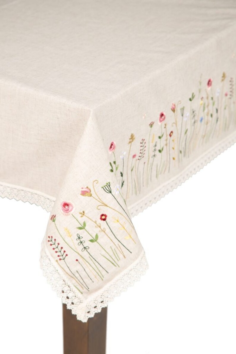 May include: A cream-colored linen tablecloth with embroidered wildflowers in pink, red, yellow, and green. The edge is finished with a delicate white lace trim. The embroidered floral design adds a touch of rustic charm to the table setting.