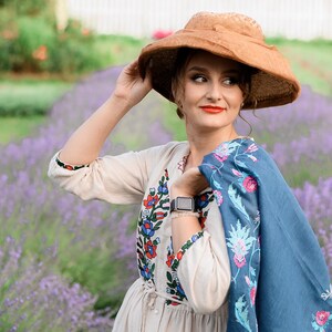 Pode incluir: Uma mulher usando um chap&eacute;u de palha, um vestido branco com bordados coloridos e um xale azul com bordados florais est&aacute; em um campo de lavanda.