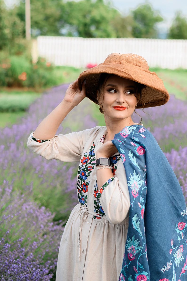 Pode incluir: Uma mulher usando um chap&eacute;u de palha, um vestido branco com bordados coloridos e um xale azul com bordados florais est&aacute; em um campo de lavanda.