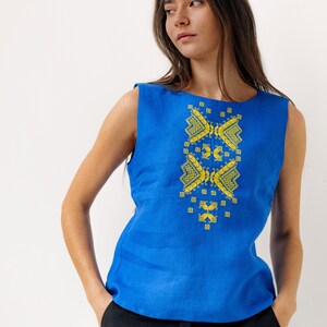 May include: A woman wearing a sleeveless blue linen top with yellow embroidered geometric patterns. She is wearing black trousers and has her hair down.