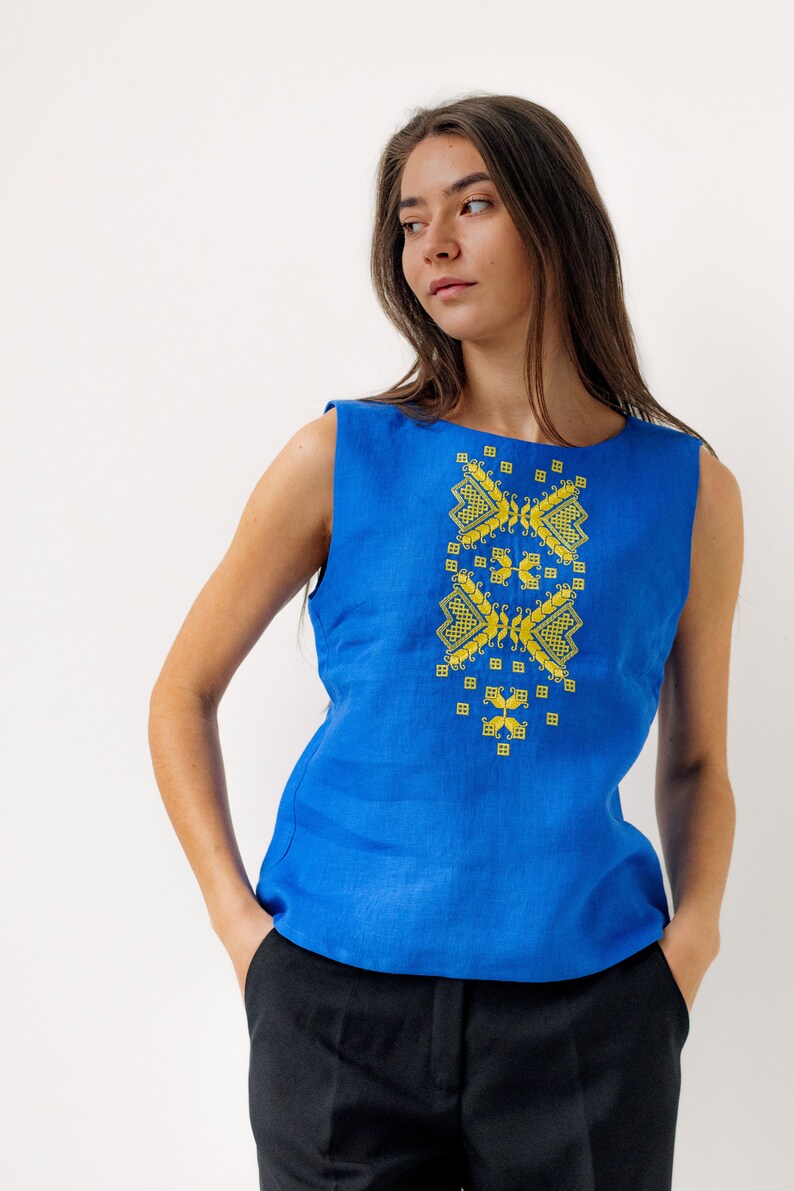 May include: A woman wearing a sleeveless blue linen top with yellow embroidered geometric patterns. She is wearing black trousers and has her hair down.