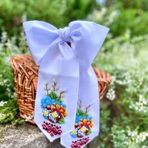 May include: A white satin ribbon with a bow, decorated with embroidered Easter eggs, flowers, and willow branches. The ribbon is tied around a wicker basket.