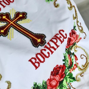 K&ouml;nnte beinhalten: Wei&szlig;es besticktes Tuch mit einem Kreuz und dem Wort "BOCKPEC" in Rot. Das Kreuz ist braun mit gelben und schwarzen Akzenten. Florale und goldene Perlenmuster umrahmen den Text und das Kreuz.