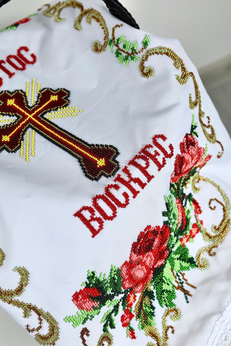 K&ouml;nnte beinhalten: Wei&szlig;es besticktes Tuch mit einem Kreuz und dem Wort "BOCKPEC" in Rot. Das Kreuz ist braun mit gelben und schwarzen Akzenten. Florale und goldene Perlenmuster umrahmen den Text und das Kreuz.