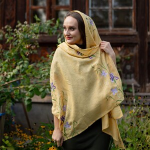 May include: A light yellow shawl with embroidered purple flowers and green stems. The shawl is draped over the shoulders and head. The background includes greenery and a wooden structure.
